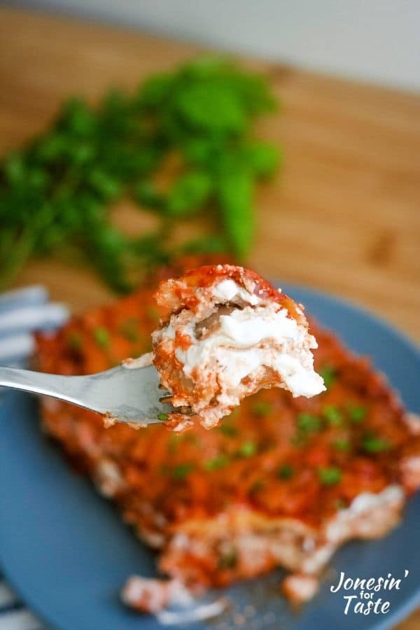 a bite of lasagna on a fork with the plate of lasagna and a bunch of fresh herbs in the background