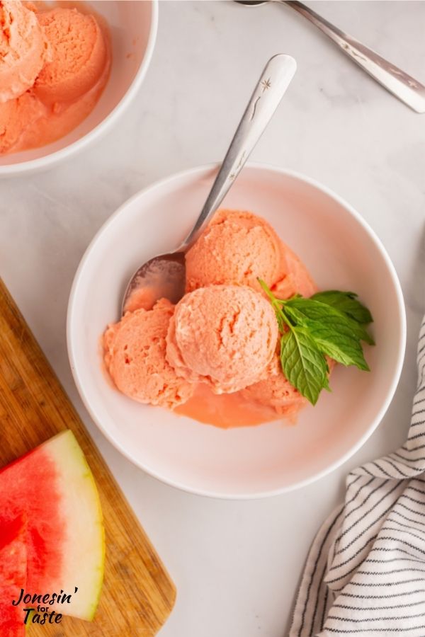 scoops of watermelon ice cream in a white bowl with a spoon on a white table next to a striped napkin and a cutting board with watermelon slices