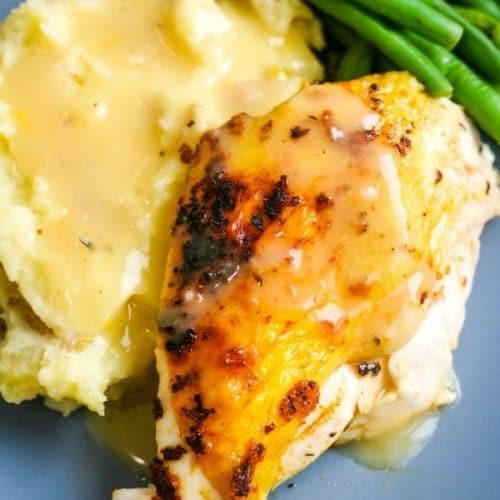 A blue plate with a piece of skin on chicken breasts sits next to a pile of green beans and on top of a scoop of mashed potatoes and both are covered in gravy