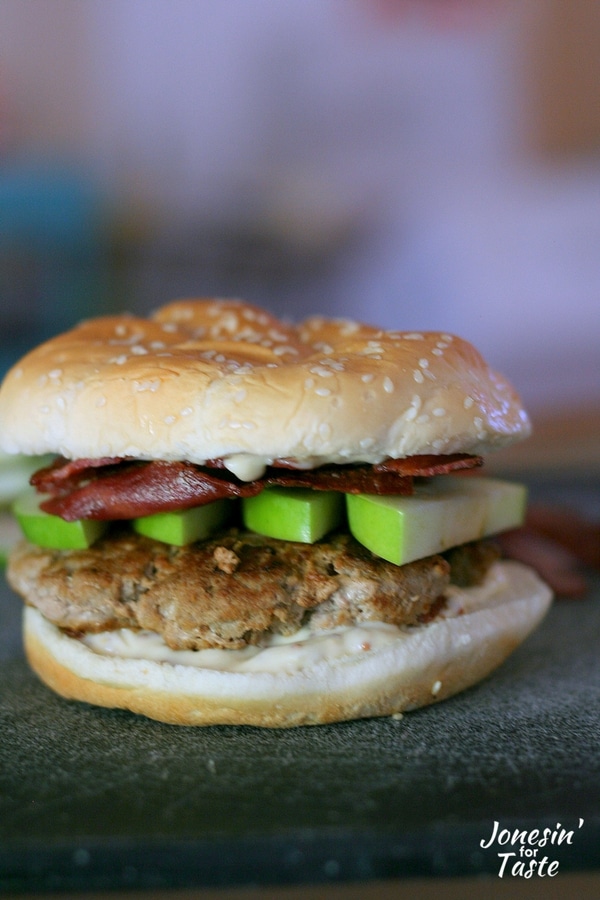 A hamburger bun with a turkey burger topped with green apple slices, turkey bacon, and honey mustard spread on a black cutting board.