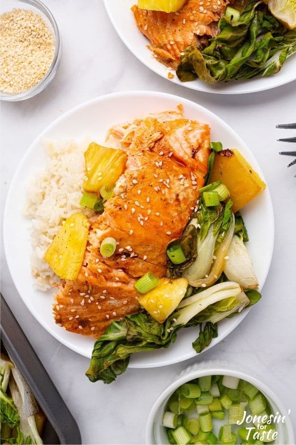 A piece of teriyaki salmon on a plate with rice, pineapple chunks, and cooked baby bok choy