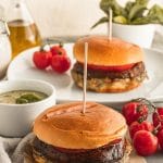 a Portobello burger sits on torn piece of parchment paper in front of a Portobello burger on a plate with various bowls of pesto and basil and tomatoes on the vine around them