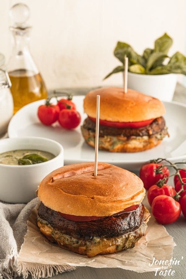 a Portobello burger sits on torn piece of parchment paper in front of a Portobello burger on a plate with various bowls of pesto and basil and tomatoes on the vine around them