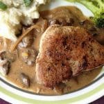 A pork chop with mushroom gravy lying on the right side of a pool of mushroom gravy with visible slices of mushrooms and onions in the gravy.