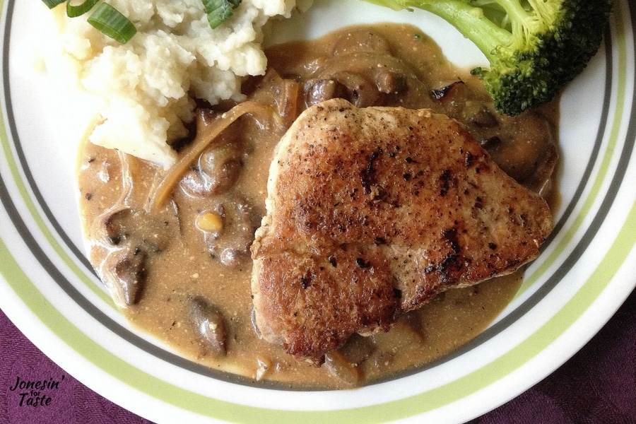 A pork chop with mushroom gravy lying on the right side of a pool of mushroom gravy with visible slices of mushrooms and onions in the gravy.