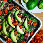 a pan of enfrijoladas topped with avocados, cilantro, and sliced tomatoes