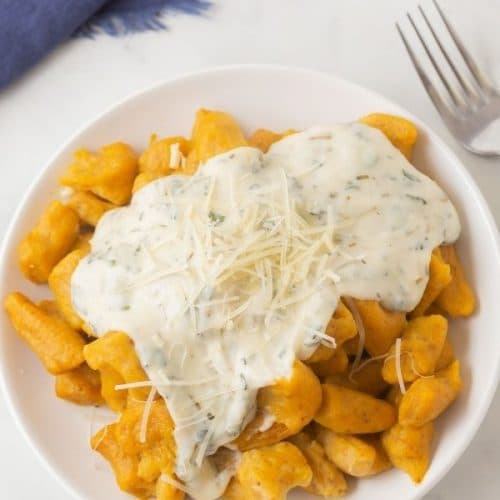 A small white plate with pumpkin gnocchi covered in a green speckled white sauce topped with freshly shredded parmesan cheese