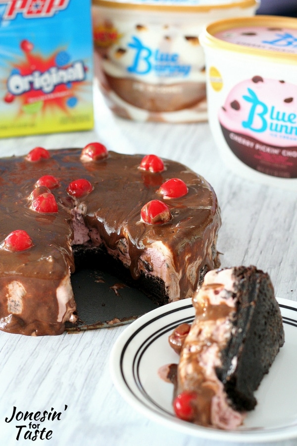 A slice of Cherry Chocolate Ice Cream Brownie Cake on a plate in front of the cake with Blue Bunny ice cream containers and Bomb Pops in the background.