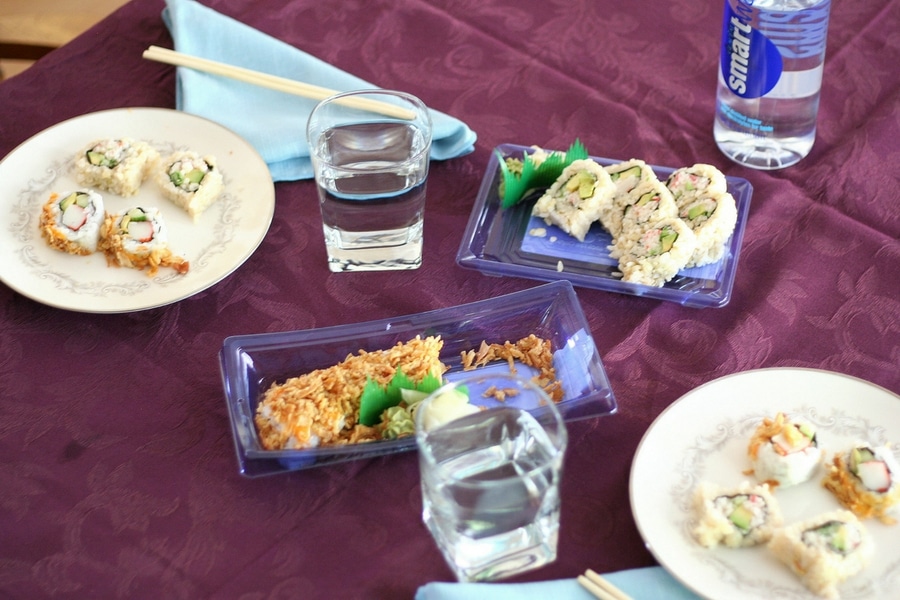 Containers of sushi, 2 plates with sushi, 2 glasses of water with blue napkins and chopsticks on a purple tablecloth