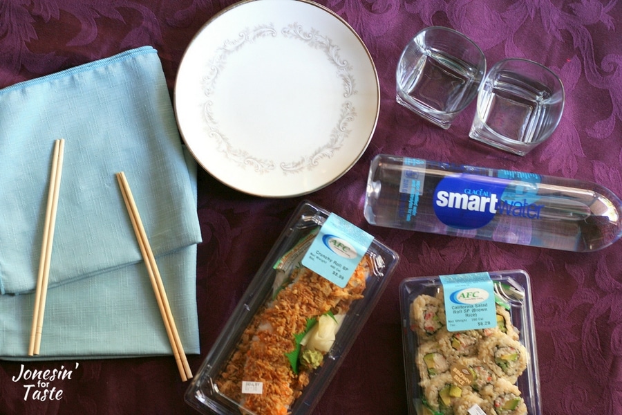 Cloth napkins, chopsticks, china plates, glasses, smartwater, and containers of sushi on a purple tablecloth