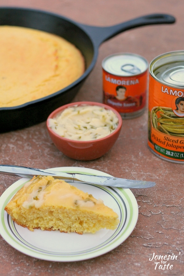 A slice of cornbread spread with jalapeno honey butter in front of a bowl, skillet of cornbread, and 2 cans