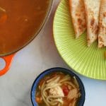 A small bowl of salsa soup next to a green plate with triangles of quesadilla and a large pot of soup next to it.