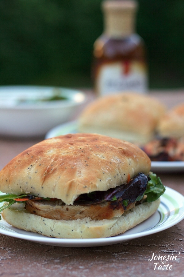 An easy Sesame grilled chicken sandwich on a plate with a plate of rolls and chicken, a bowl of salad, and a bottle of marinade behind it.