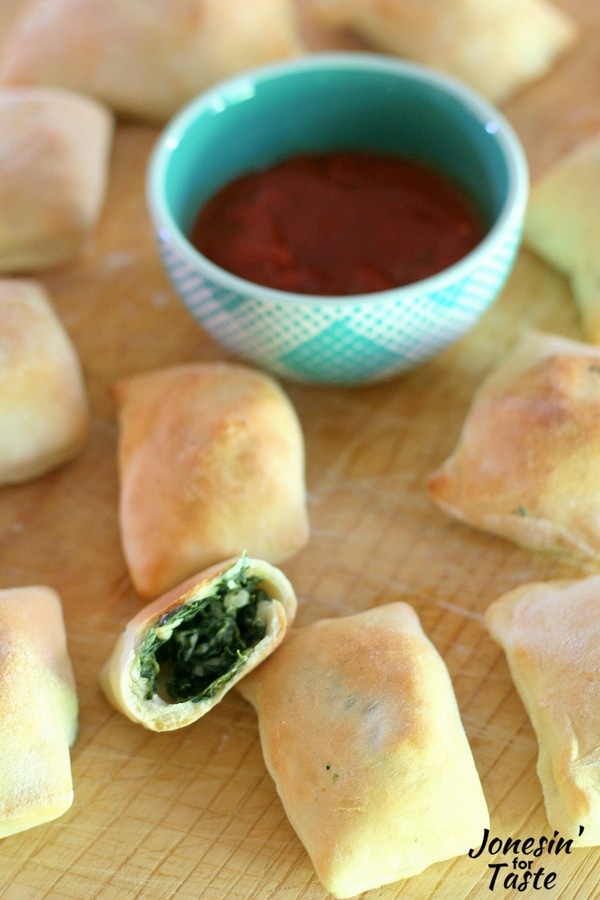 A batch of Mini Spinach Calzones on a cutting board, with one ripped in half with a bowl of dipping sauce.