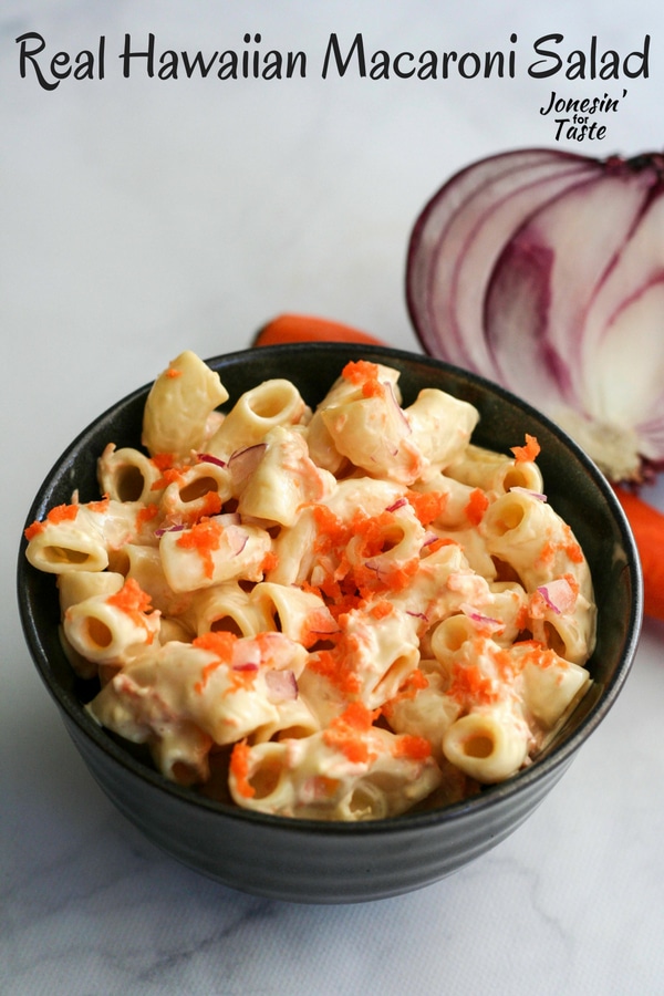 A black bowl full of hawaiian macaroni salad sitting in front of half a red onion and a carrot