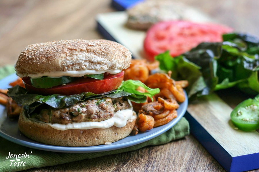 A plate with a spicy jalapeno black pepper turkey burger with sweet potato fries and a wooden board with lettuce, tomato, and additional turkey burgers in the background.