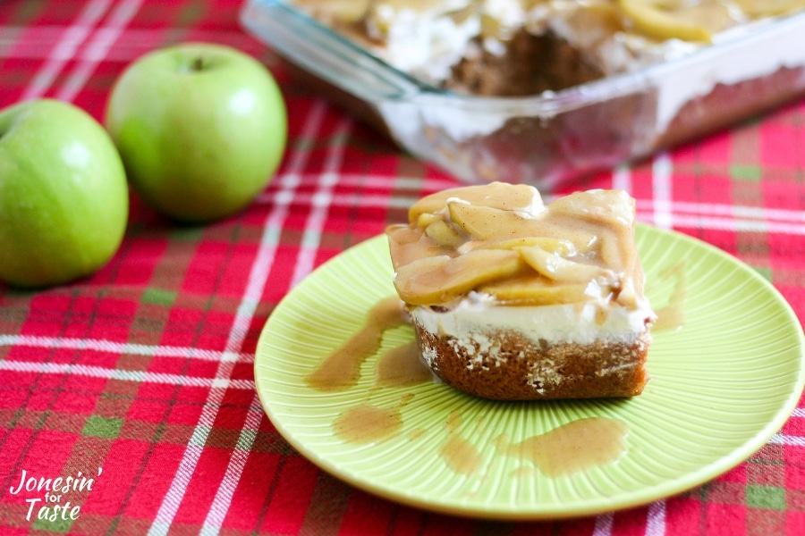 A slice of apple spice poke cake on a green plate next to the cake pan and two apples