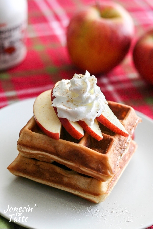 A stack of chai spiced apple waffles with whipped cream next to whole apples and maple syrup