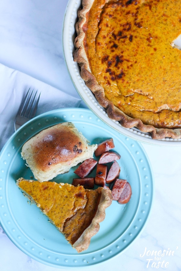 A slice of pumpkin quiche on a blue plate with a roll and smoked sausage