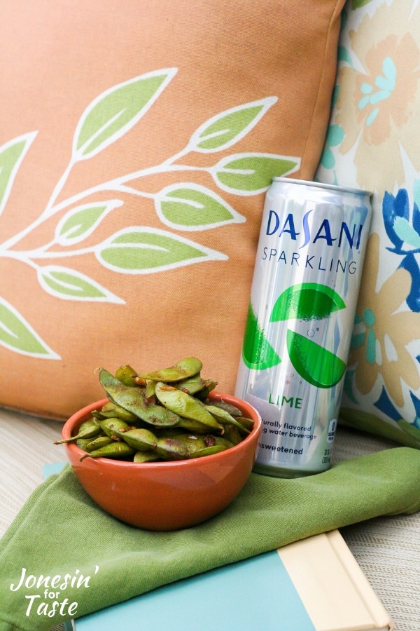 A bowl of garlic soy edamame next to a can of sparkling water and pillows