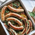 a white casserole dish filled with sliced potatoes, bacon, herbs, and browned bratwurst sausages
