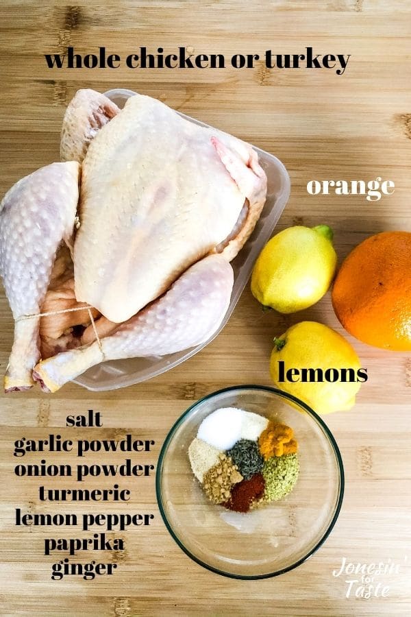 a raw whole chicken, 2 lemons, an orange, and a bowl of spices sit on a wooden background