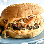a burger sits on a blue plate. It is spilling over the bun with ingredients like an orange hued sauce and a coleslaw