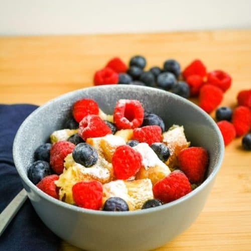 a bowl of golden yellow cubes of bread sit in a grey blue bowl topped with a dusting of powdered sugar and fresh berries