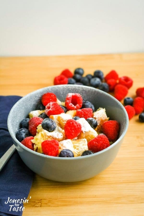 a bowl of golden yellow cubes of bread sit in a grey blue bowl topped with a dusting of powdered sugar and fresh berries
