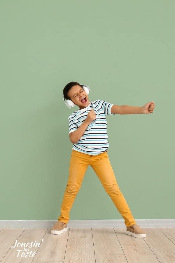 a young lighter skinned black boy with brown hair in a blue and white striped shirt, yellow pants, and headphones is listening to music and dancing while pretends to sing into a mike