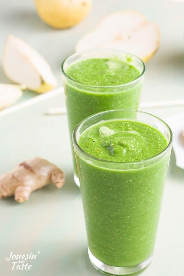 2 tall glasses filled to the top with a vibrant green smoothie. Cut pears, ginger root, and straws are strewn about around the glasses.