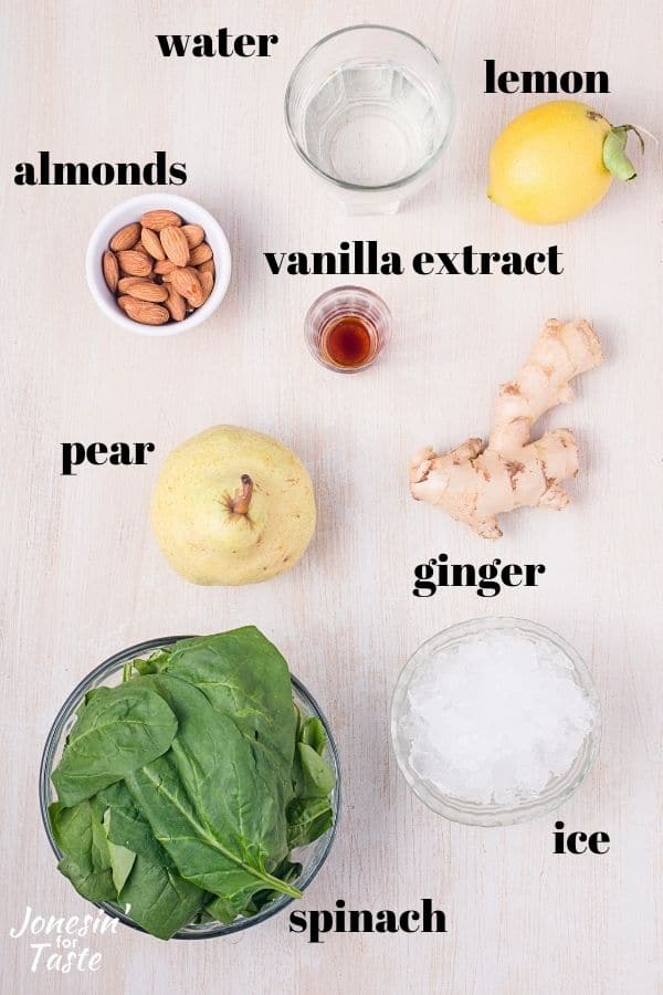 a glass of water, a lemon, ginger root, a bowl of ice, spinach, a pear, and bowls of vanilla extract and almonds sit on a light background