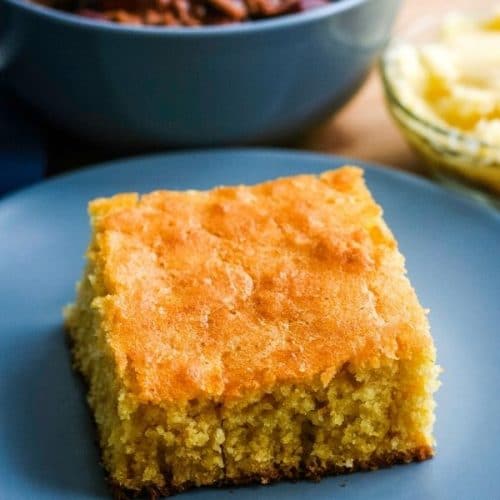 a square slice of golden brown cornbread on a blue plate in front of a bowl of chili