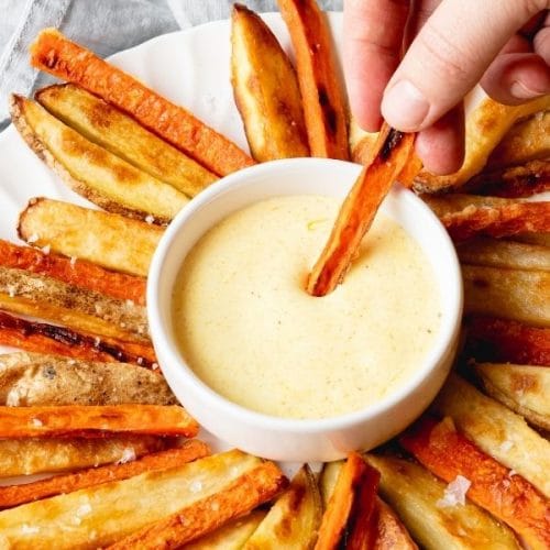 you see a person's hand is dipping a carrot fry into a small white bowl with a light yellow curry mayo dipping sauce