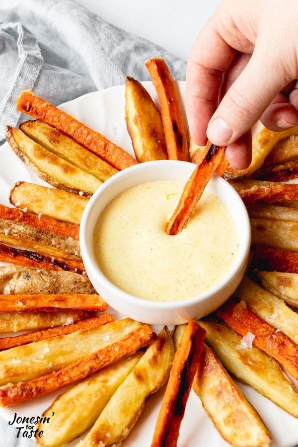 you see a person's hand is dipping a carrot fry into a small white bowl with a light yellow curry mayo dipping sauce