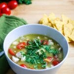 a bowl of green chicken chili on a wooden backdrop surrounded by chips, a green napkin, cherry tomatoes, and a bunch of cilantro