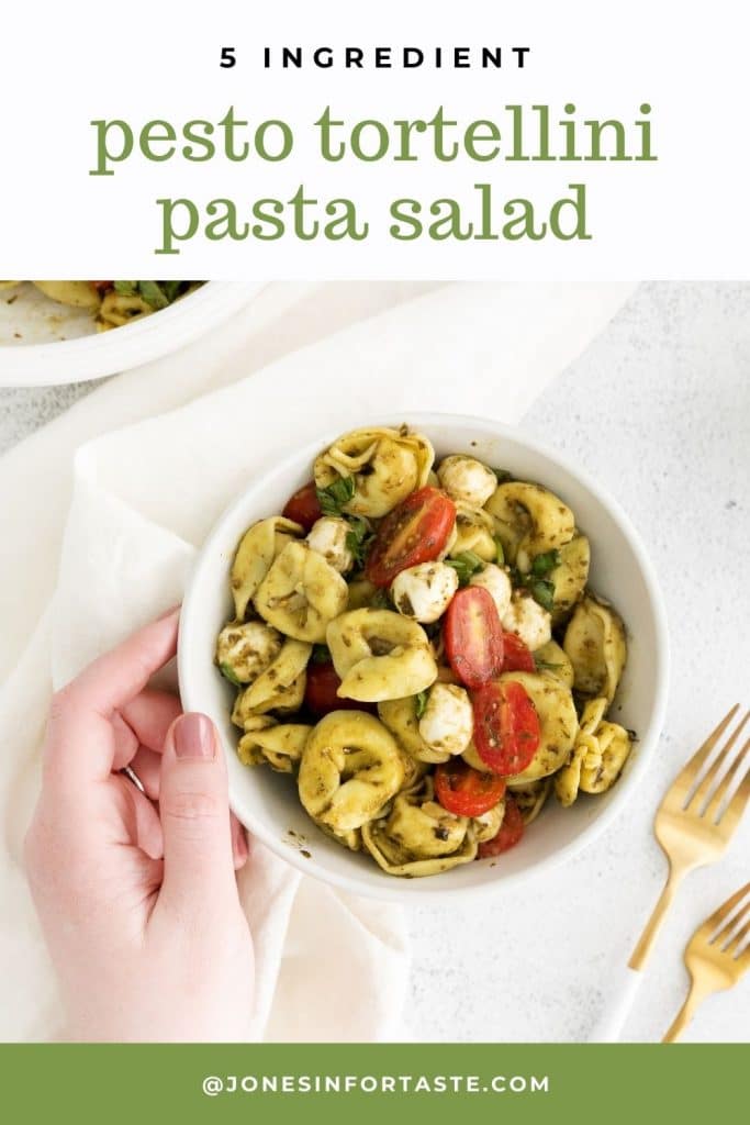 a hand softly holding a white bowl of pesto tortellini pasta salad