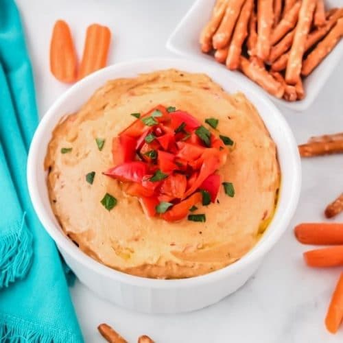 a large white bowl with creamy red pepper hummus topped with chopped red peppers and sprinkled with chopped parsley surrounded by carrot sticks and pretzel sticks