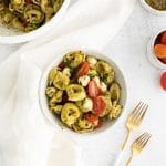 a portion of tortellini pasta salad in a white bowl surrounded by a white linen napkin, white handled forks, and other various bowls with the pasta salad and various ingredients.