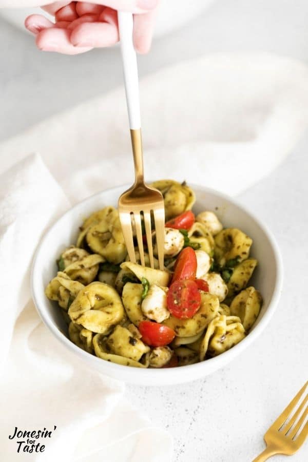 a hand holding a fork. The fork is pressing down on a tortellini in a white bowl to pick up.