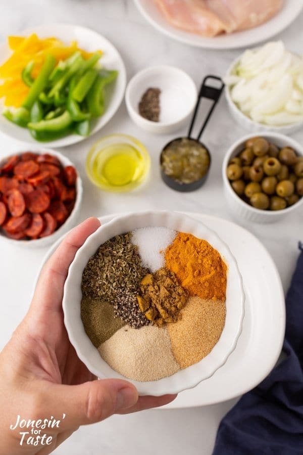 a bowl of spices in small little mounds inside the bowl is held above a work surface with other ingredients laid out on it
