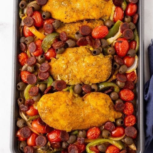 a cookie sheet filled with chicken breasts coated in seasoning and a glaze surrounded by multicolored tomatoes, chorizo, olives, peppers and onions.