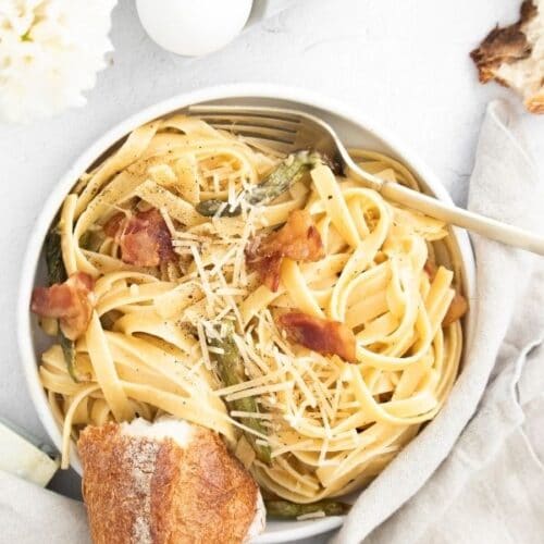 a top down view of Fettucine carbonara with bacon and roasted asparagus in a white dish on white linens.